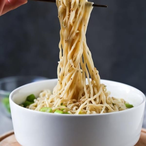 Chopsticks lifting seasoned noodles with sesame seeds and green vegetables from a white bowl.
