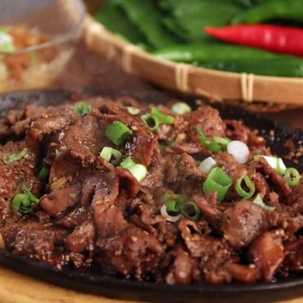 Sizzling bulgogi on a black plate, garnished with green onions and sesame seeds, with fresh vegetables in background.